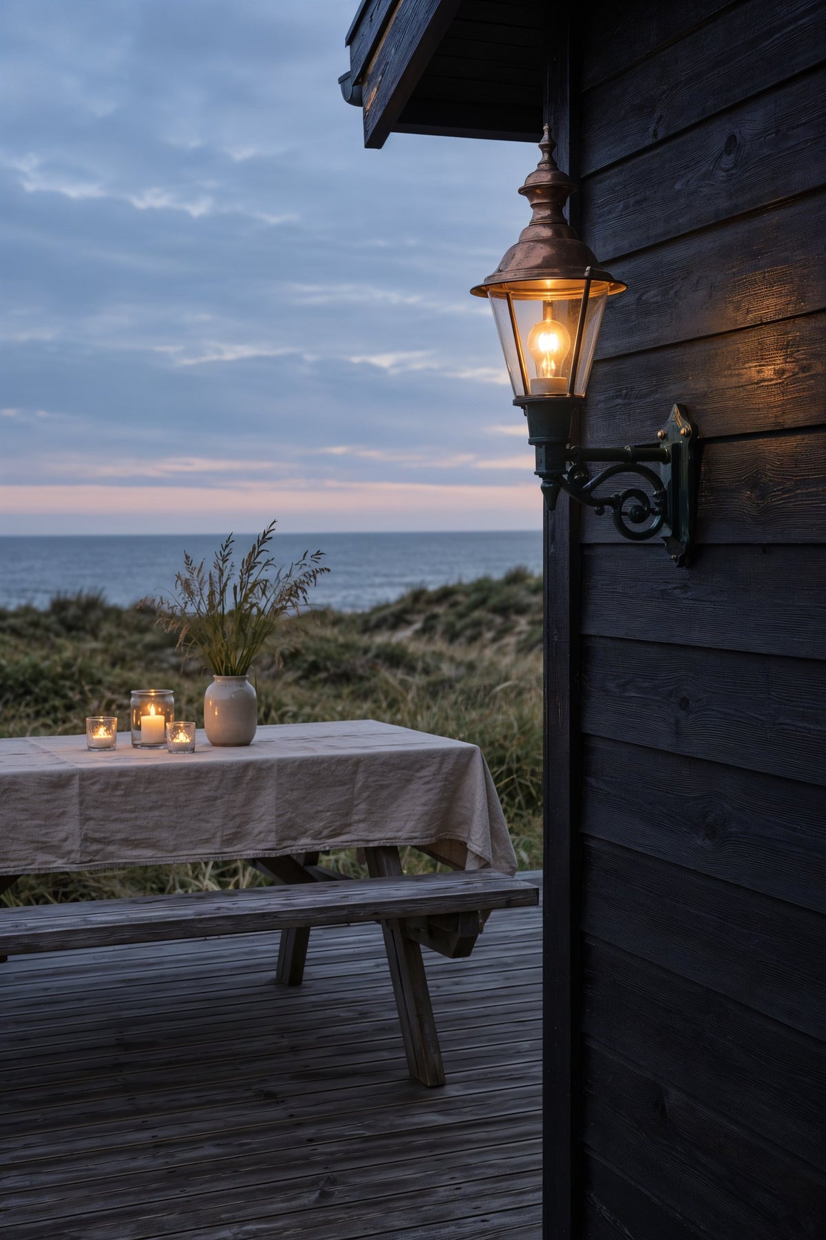 Sort træsommerhus ved klitterne med væglampe i kobber over terrassen